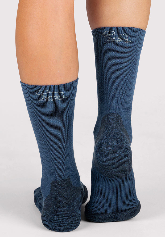 A person wearing Woolx Womens Harbor Crew Socks in Majolica Blue, featuring darker blue heels and white text near the top back, stands on a light surface.
