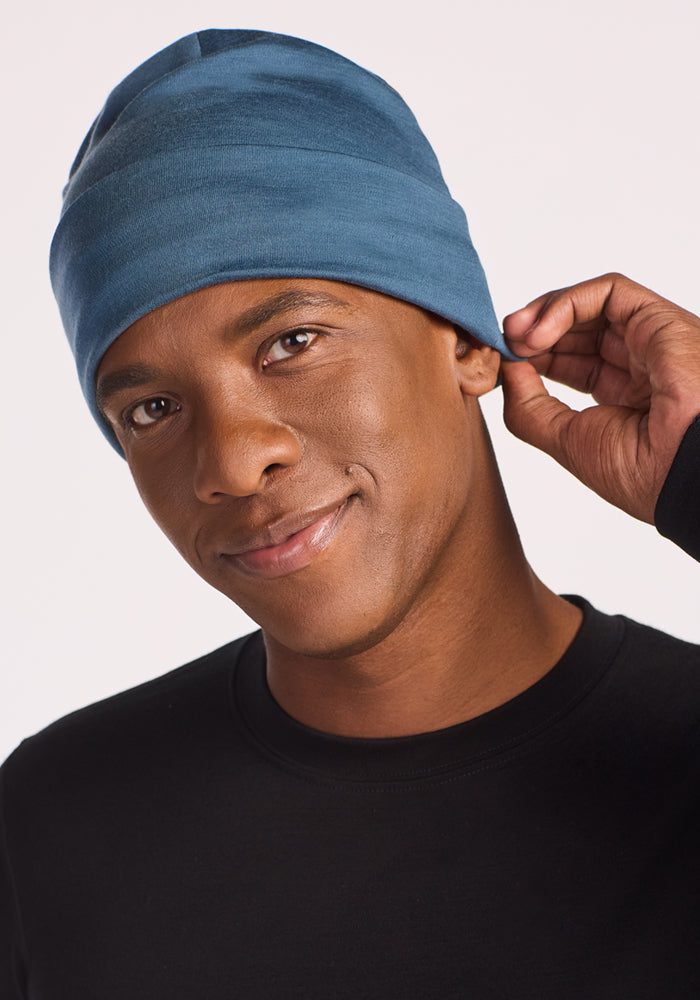 A person in a black shirt smiles at the camera while adjusting their Woolx Journey Hat in Steel Blue. The plain, light background highlights the hat's soft Merino wool and moisture-wicking comfort.