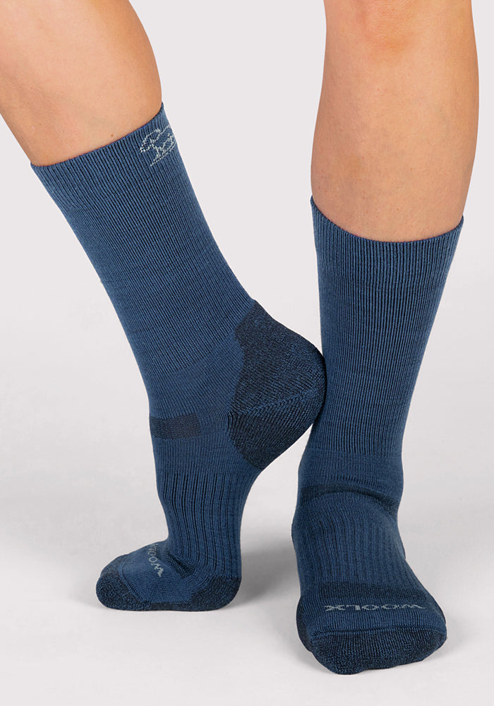 A person models the Woolx Womens Harbor Crew Sock in Majolica Blue, standing on a light gray surface with one foot flat and the other raised to display the socks from different angles.