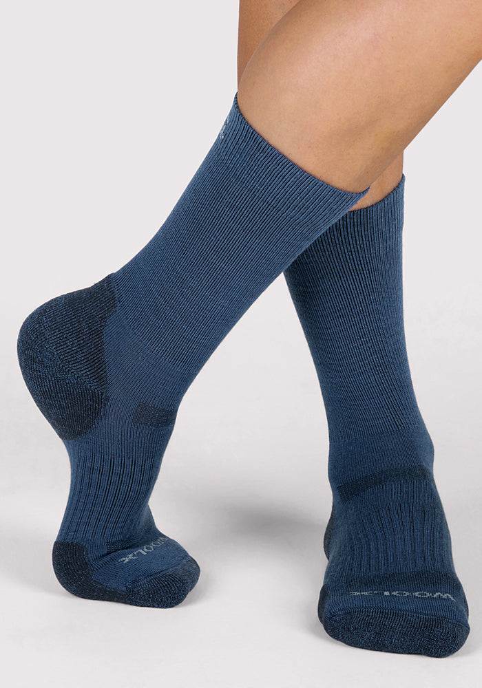 A person is wearing Woolx Womens Harbor Crew Socks in Majolica Blue. Their feet are crossed at the ankles against a plain light gray background.