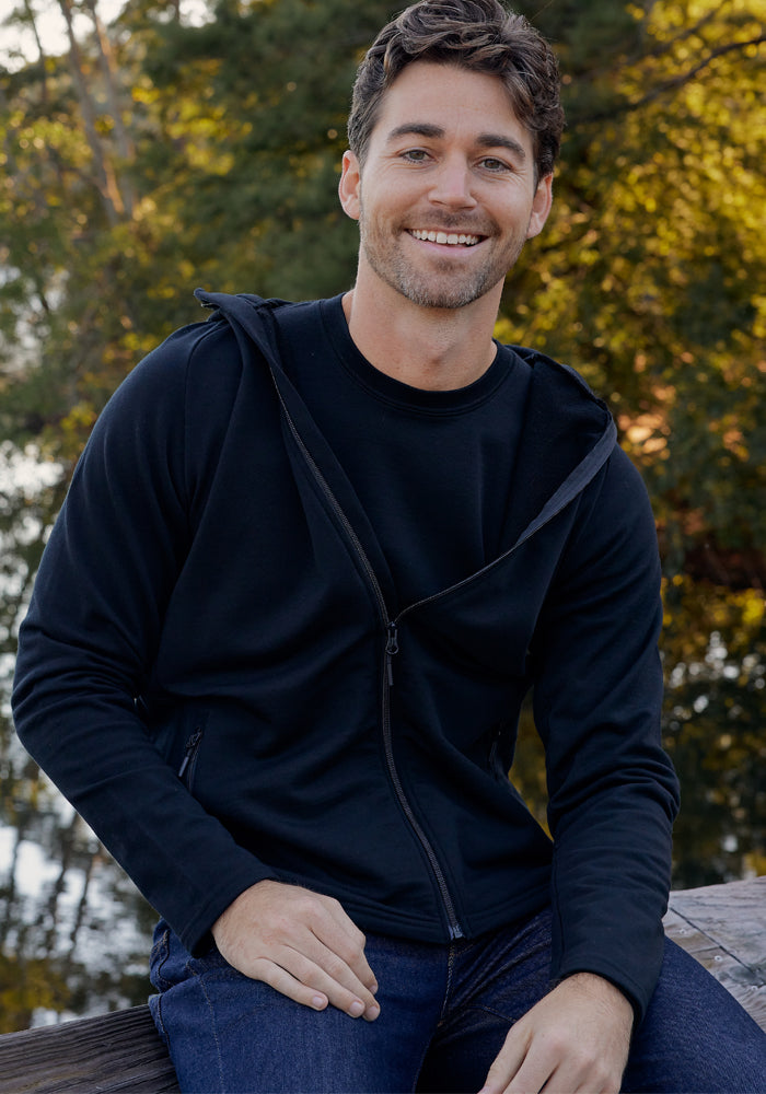 A man with short brown hair and a beard sits outdoors on a wooden surface, smiling at the camera, wearing the Woolx Boulder Hoodie in black and blue jeans, with trees and sunlight in the background.
