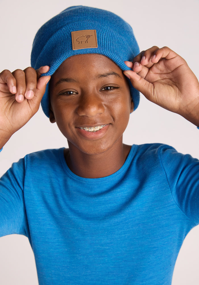 A smiling child in a blue long-sleeve shirt wears the Woolx Kids Baylor Beanie - Blue Razz, featuring a brown patch, and holds the edges of this cozy winter hat with both hands.