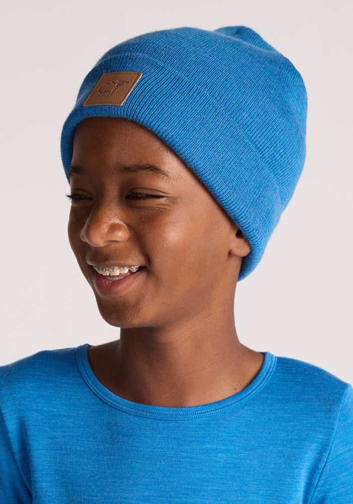 A smiling child in a Woolx Kids Baylor Beanie - Blue Razz, paired with a matching blue shirt, looks slightly to the side against a plain light background.