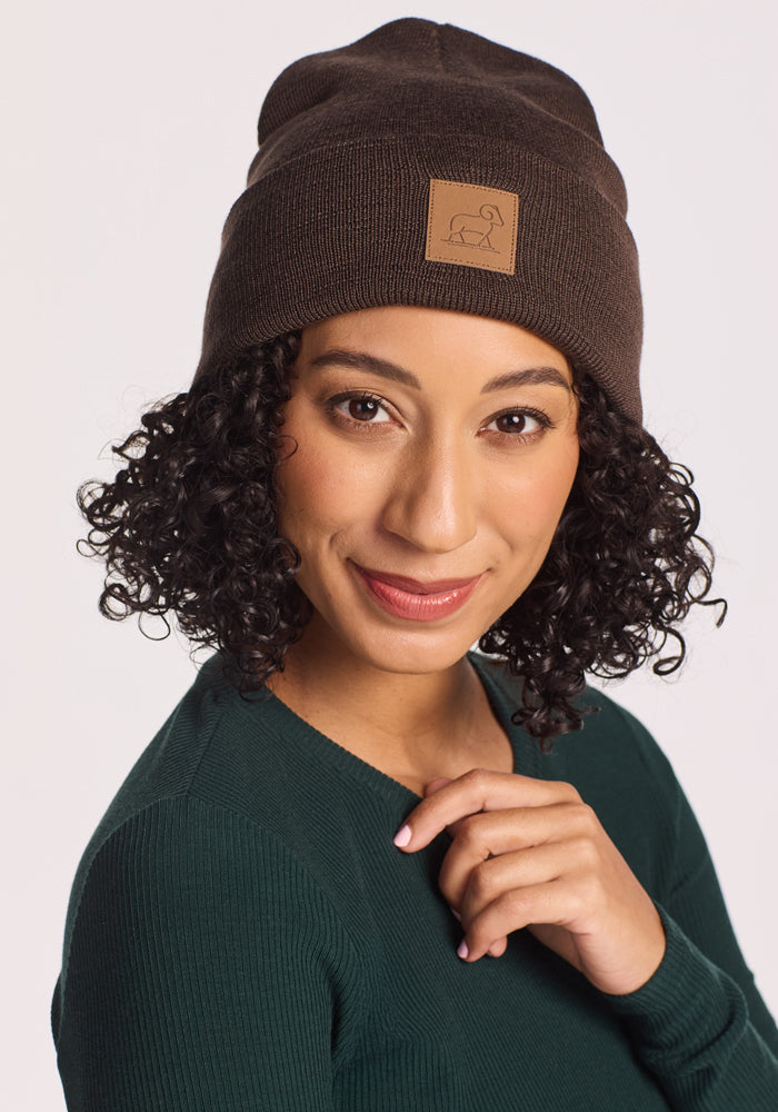 A woman with curly hair wears the Woolx Baylor Beanie in Bracken—a dark brown merino wool hat with a small dog logo patch. She smiles softly at the camera, dressed in a dark green top and touching the beanie.