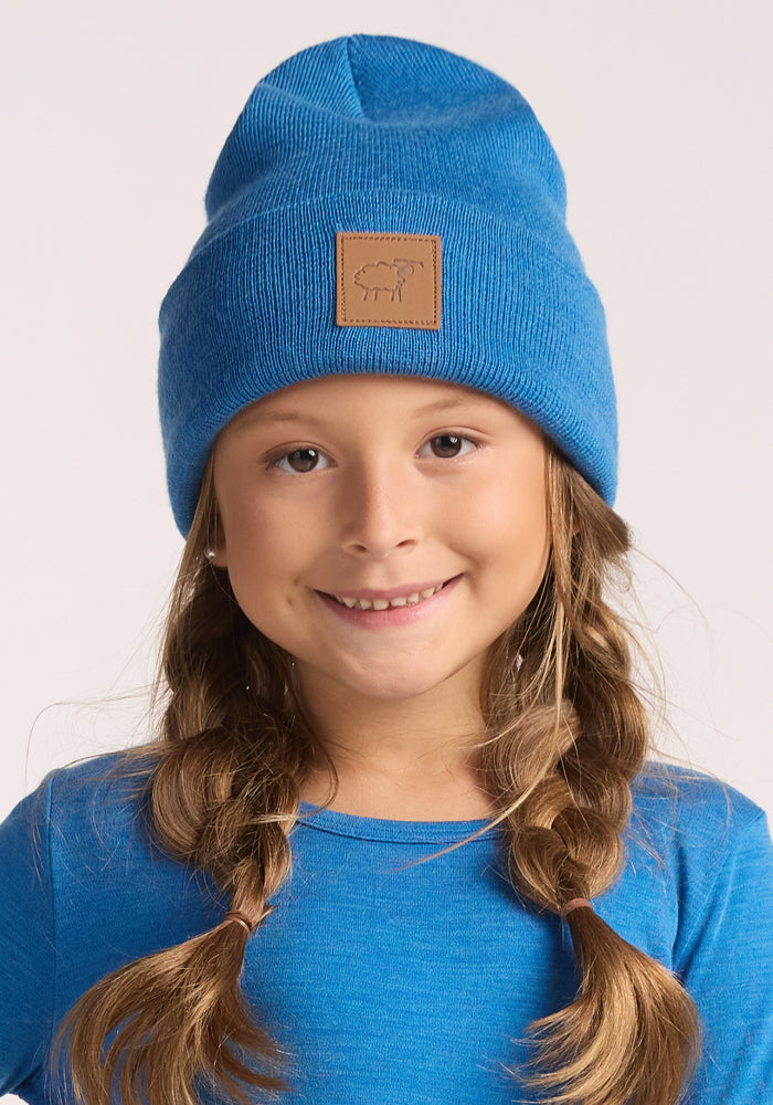 A smiling girl with long brown braided hair is wearing the Woolx Kids Baylor Beanie in Blue Razz, which features a brown patch with a sheep logo—a perfect winter hat for kids.