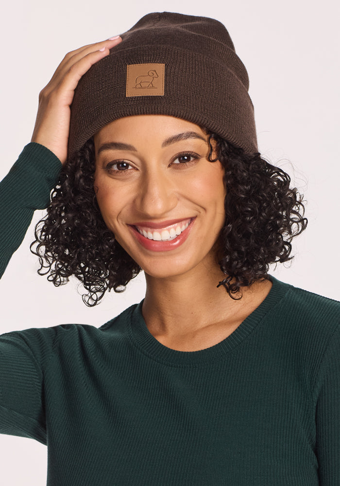 A smiling person with curly hair wears the Woolx Baylor Beanie in Bracken—a dark brown merino wool winter hat with a patch—paired with a green long-sleeve shirt, touching the beanie against a light background.