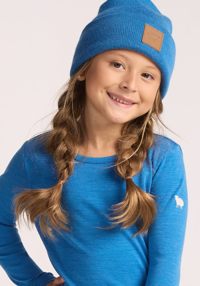 A young girl with light brown braids smiles, wearing the Woolx Kids Baylor Beanie in Blue Razz and a matching blue shirt featuring a small white sheep logo on the sleeve.
