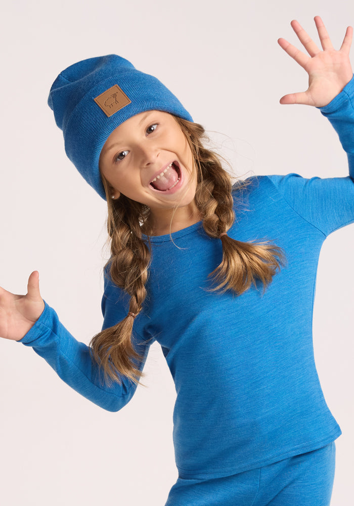 A young girl with braided hair smiles widely and raises her hands playfully while wearing the Woolx Kids Baylor Beanie in Blue Razz, paired with a matching long-sleeve shirt against a plain light background.