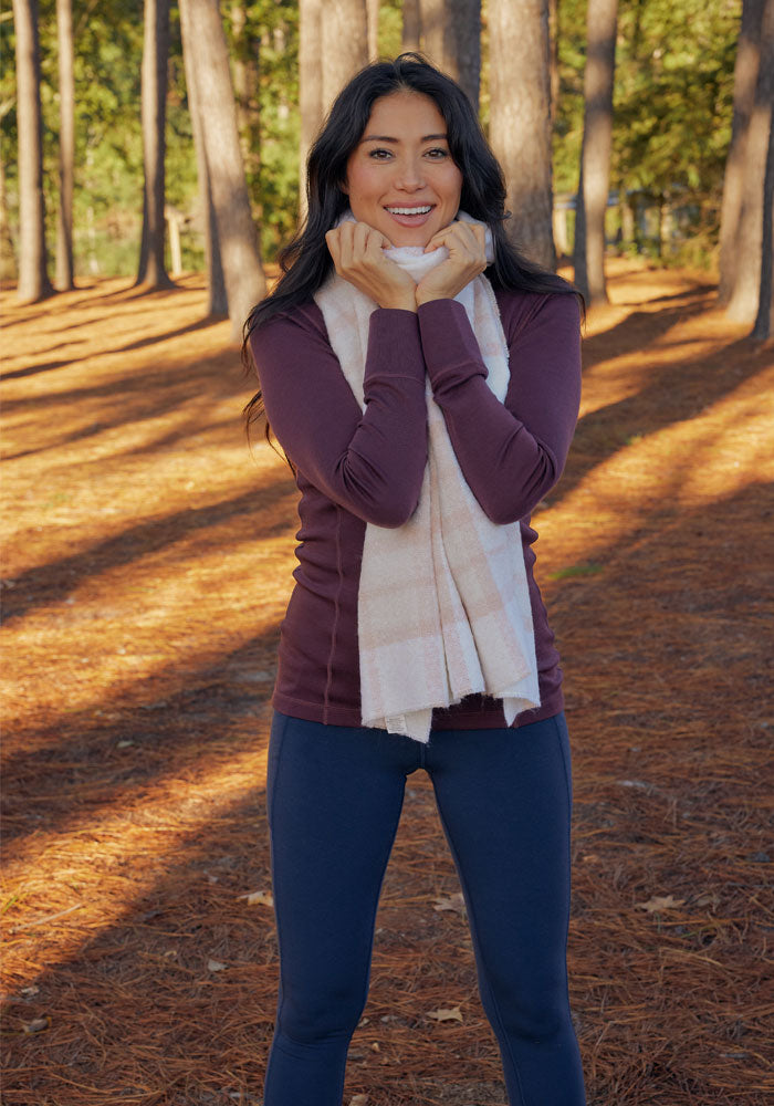 A woman stands outdoors among tall trees, smiling with her hands under her chin. She wears a maroon long-sleeve top, navy leggings, and the Woolx Bradie Scarf in Mauve Plaid. Sunlight filters through the trees, casting shadows on the ground.
