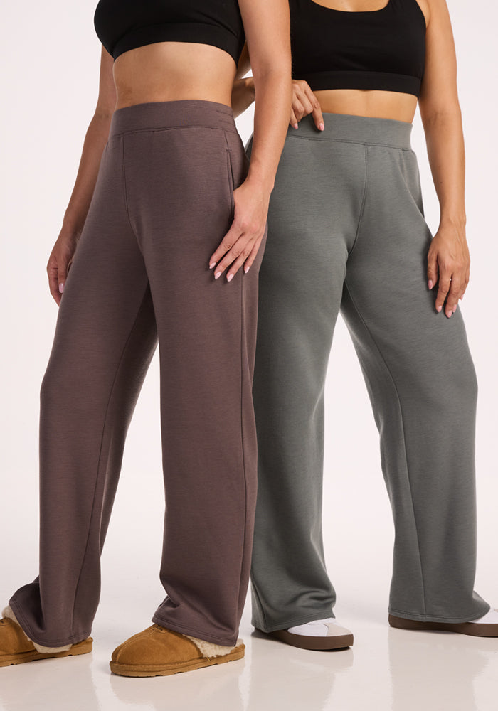 Two women stand side by side, faces out of view, wearing relaxed fit black crop tops and Woolx Bree Lounge Pants in Sedona Sage—one paired with matching slippers, the other with white slippers—set against a plain white background.