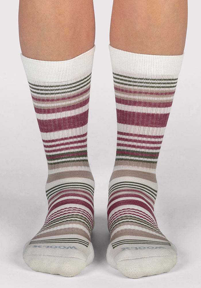 A pair of feet wear Woolx Womens Everyday Crew Socks in Buttercream Stripe—moisture-wicking, white cushioned crew socks with burgundy, green, beige, and brown stripes—standing on a light surface, with bare legs above the mid-calf socks.