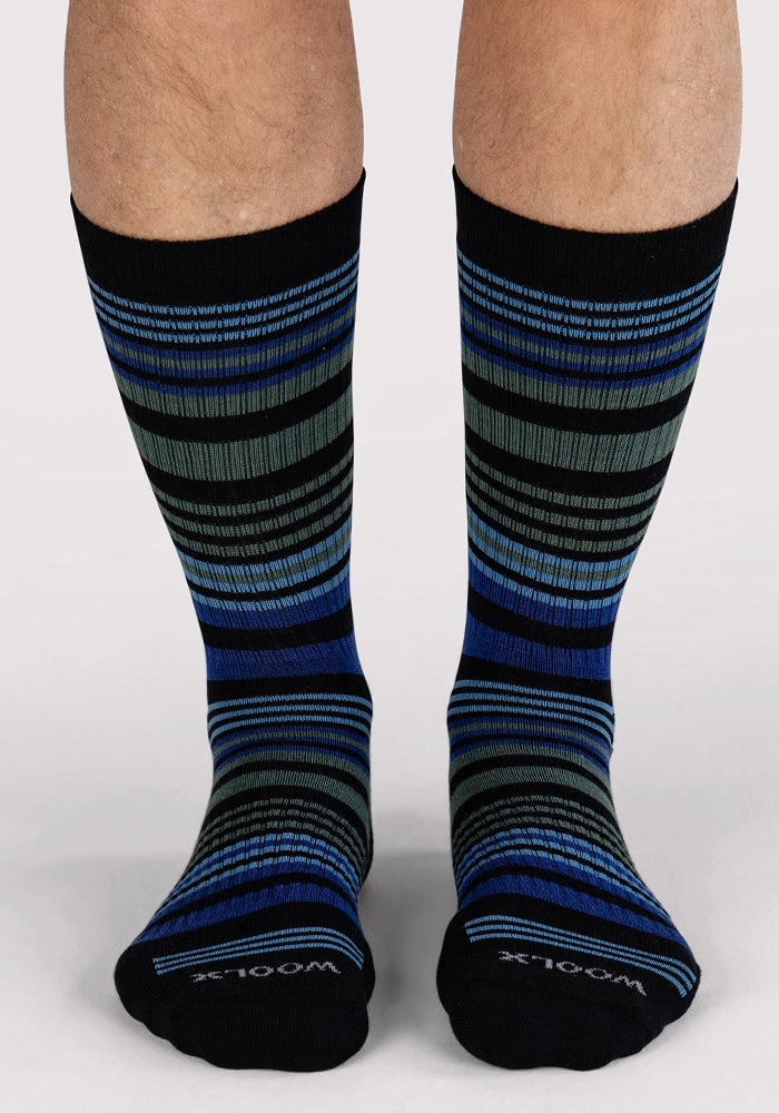 A person stands on a light surface wearing Woolx Mens Everyday Crew Socks in Black Stripe, featuring blue, gray, and green stripes and "WOOL" on the cushioned toes.