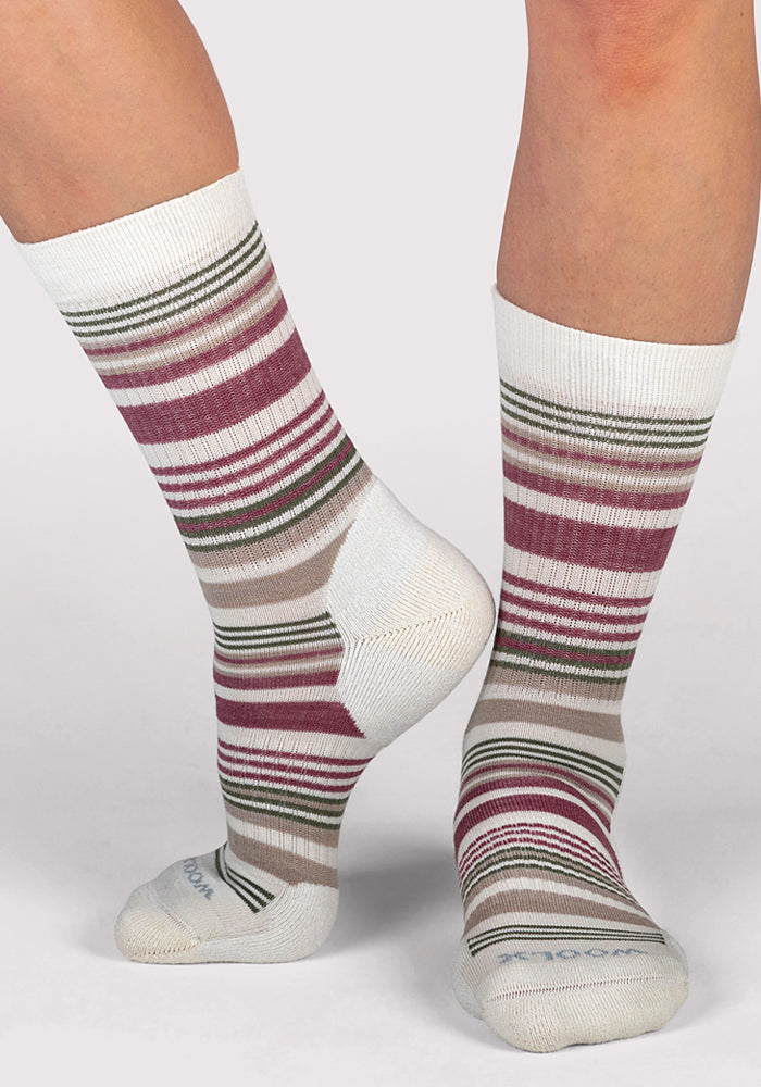 Wearing Woolx Womens Everyday Crew Socks in Buttercream Stripe, featuring moisture-wicking fabric and horizontal stripes in burgundy, green, beige, and brown, she stands on a white background with one foot flat and the other raised on her toes.