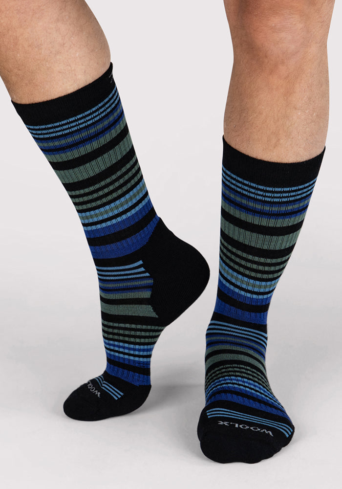 A person wears Woolx Mens Everyday Crew Socks in Black Stripe, featuring moisture-wicking fabric, horizontal black, blue, and gray stripes, and solid black toes and cuffs, standing against a light background.