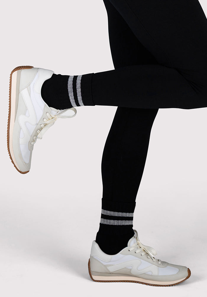 A person wears Woolx Womens Varsity Mini Crew Socks - Black Volt with black leggings and white sneakers, standing with one leg bent and balancing on the other against a plain light background.