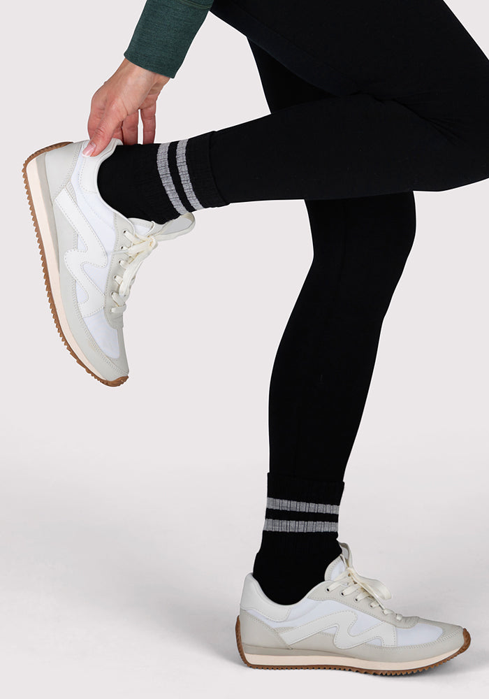 A person wearing black leggings, Woolx Womens Varsity Mini Crew Socks - Black Volt, and white sneakers stands on one leg, holding the opposite ankle against a plain background.