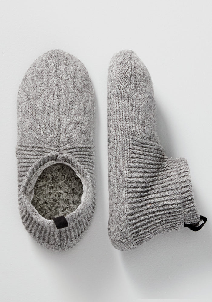The Woolx Womens Gripper Slippers in Gentle Grey are shown, featuring ribbed cuffs and a soft, textured merino wool design for warmth and comfort at home. One slipper is viewed from the top, the other from the side.
