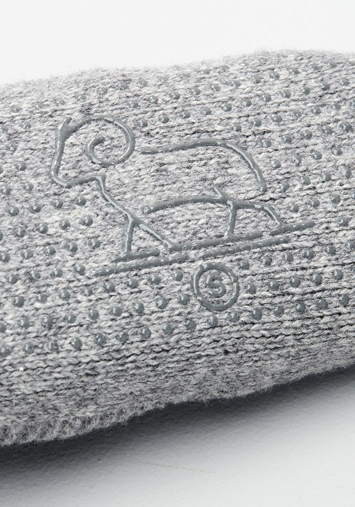 Close-up of Woolx Womens Gripper Slippers - Gentle Grey shows gray textured fabric with a dotted non-slip sole, featuring an embossed outline of a ram standing on a curved line.