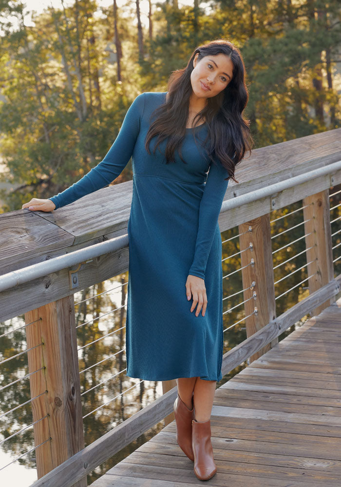 A woman with long dark hair wears the Woolx Caroline Ribbed Twirl Dress in Toasted Garnet and brown ankle boots, leaning on a wooden bridge railing with trees and sunlight in the background.