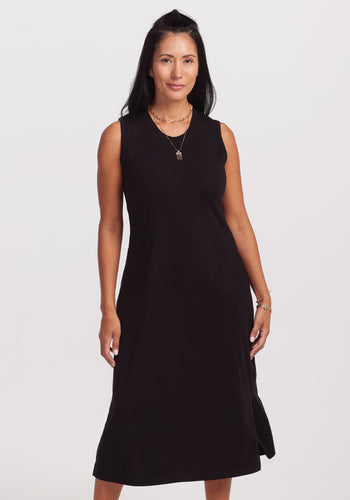A woman with long dark hair wears the sleeveless Woolx Cassie Dress in black against a plain light background. Her subtle jewelry enhances the outfit as she looks forward with a slight smile. | Denia is 5’8”, wearing a size S