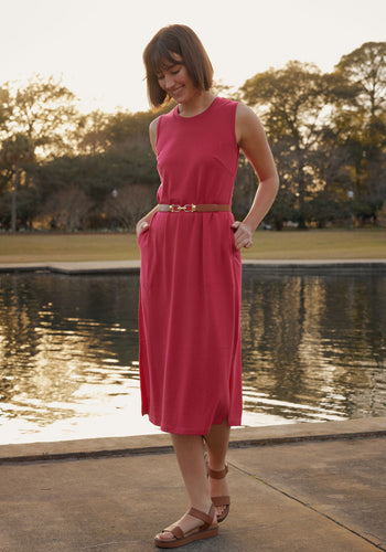 A woman in the Woolx Cassie Dress - Poppy Spark stands by a waterfront at sunset, smiling with hands in her pockets. The sleeveless merino wool dress is cinched with a brown belt as golden light reflects off the water and trees behind her.