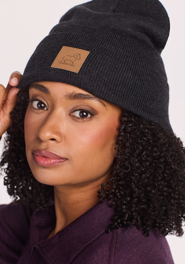 A woman with curly hair and a serious expression wears the Woolx Baylor Beanie in Carbon Black, featuring a leather patch with a simple line drawing. She pairs it with a dark purple top, gently touching the edge of her winter hat.