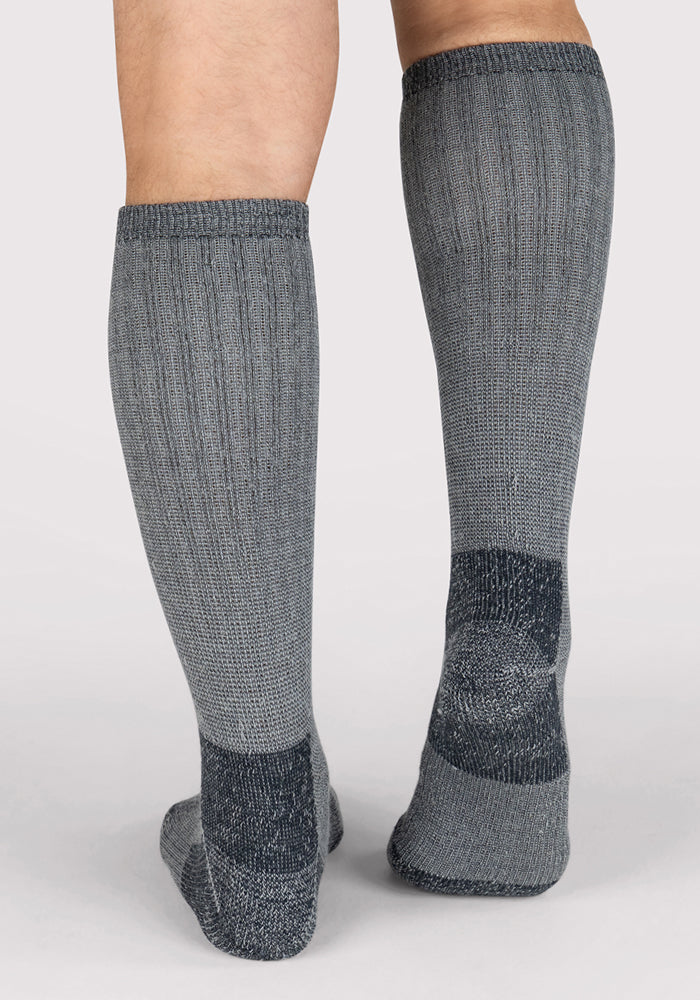 Back view of a person wearing Woolx Mens Extreme Over The Calf Socks in Charcoal Heather, heavyweight Merino wool with full cushioning and darker heels and toes, standing on a light-colored surface.