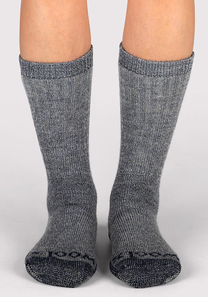 A pair of feet in Woolx Womens Extreme Crew Socks - Charcoal Heather, thick heavyweight merino wool with subtle ribbing, stand on a light gray surface. These mid-calf socks are perfect for warmth in cold weather.