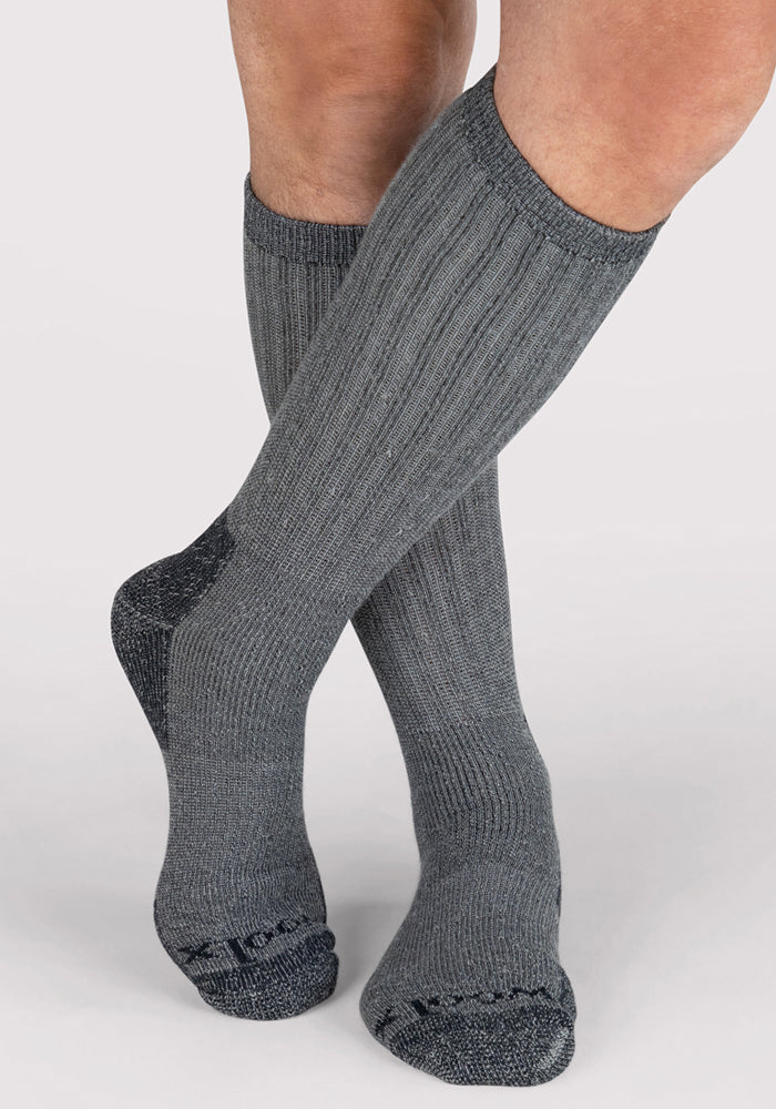 A person wearing Woolx Mens Extreme Over The Calf Socks in Charcoal Heather, with full cushioning, ribbed design, and darker gray reinforced heels and toes, stands on a light-colored background.