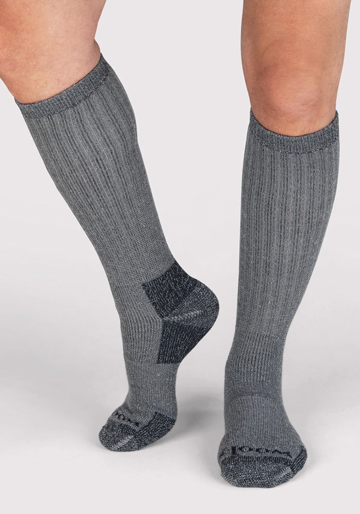 A person wearing Woolx Mens Extreme Over The Calf Socks in Charcoal Heather, featuring heavyweight Merino wool, full cushioning, and darker reinforced toes and heels, stands on a light background.