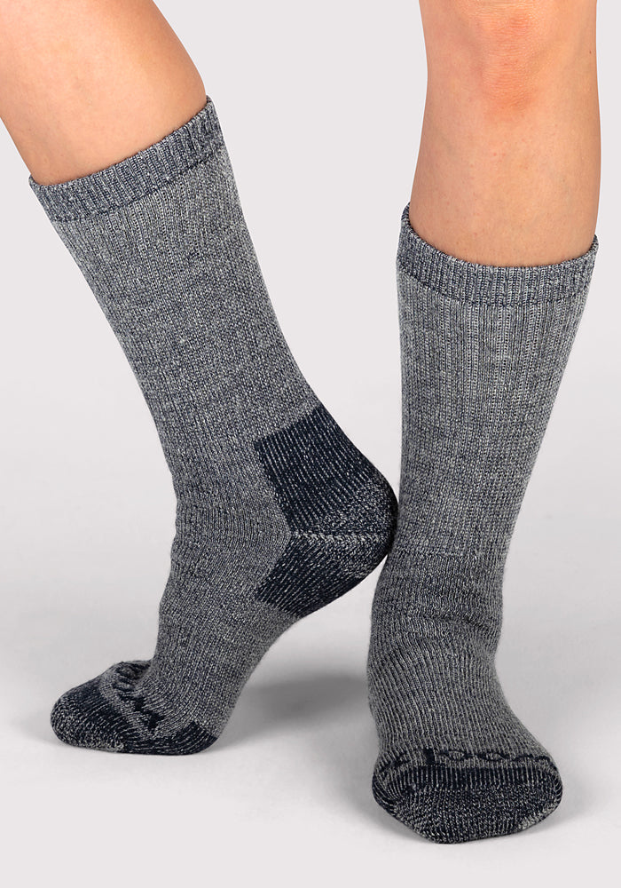 A woman wearing Woolx Womens Extreme Crew Socks in Charcoal Heather, featuring darker heels and toes, stands on a light gray background with one foot flat and the other raised—perfect winter socks for chilly days.