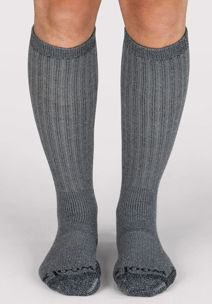 A person wearing Woolx Mens Extreme Over The Calf Socks in Charcoal Heather, with full cushioning, stands with feet flat on a plain white background.