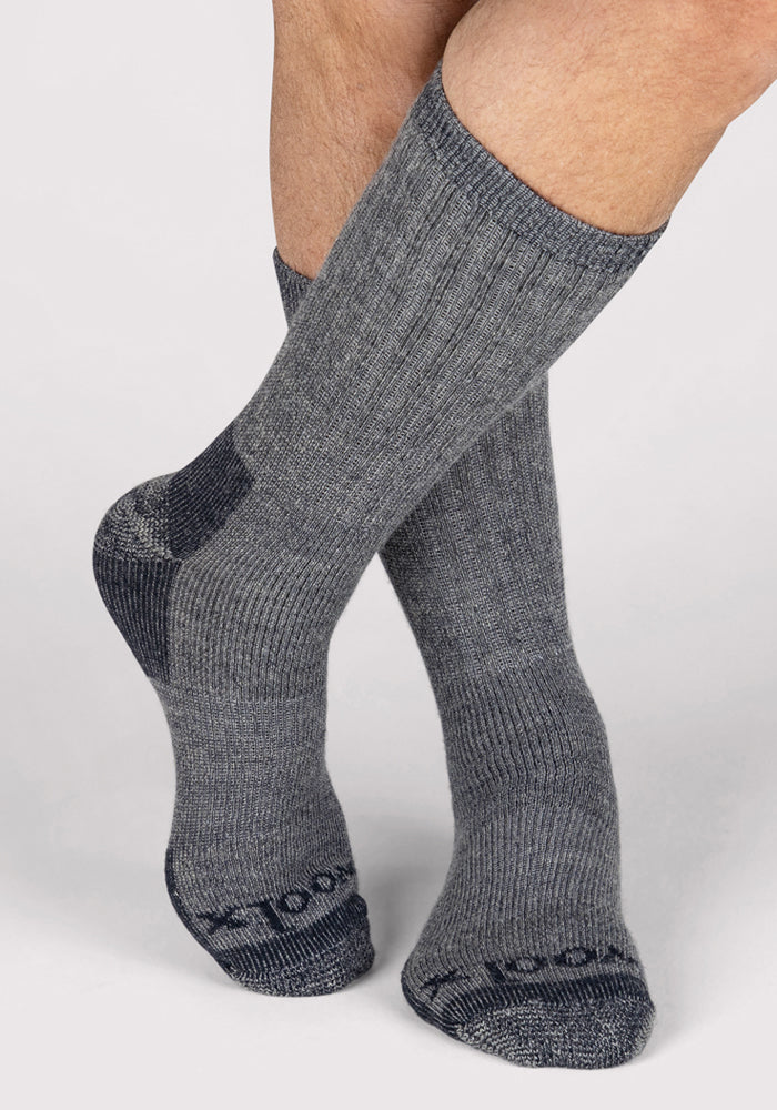 A person wearing Woolx Mens Extreme Crew Socks in Charcoal Heather, featuring darker heels and toes, stands with one foot crossed in front of the other against a light background.