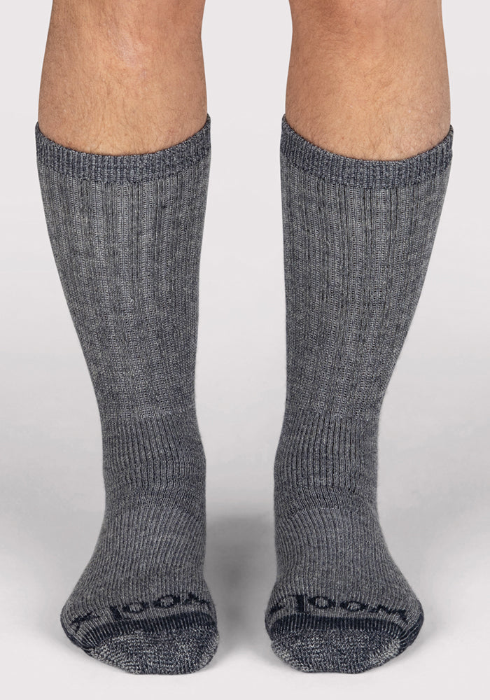 A pair of feet wearing Woolx Mens Extreme Crew Socks in Charcoal Heather, made from heavyweight merino wool for warmth and comfort, rise to mid-calf while standing on a light-colored surface.