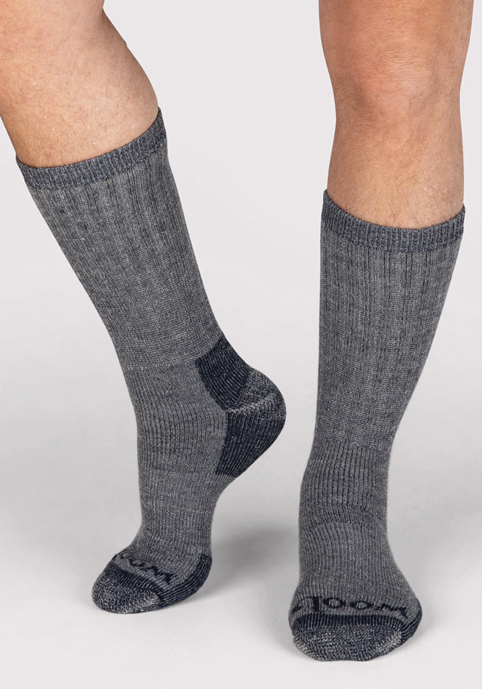 A person wears Woolx Mens Extreme Crew Socks in Charcoal Heather, standing on a light surface with one foot slightly raised, showcasing the ribbed warmth and comfort ideal for winter.