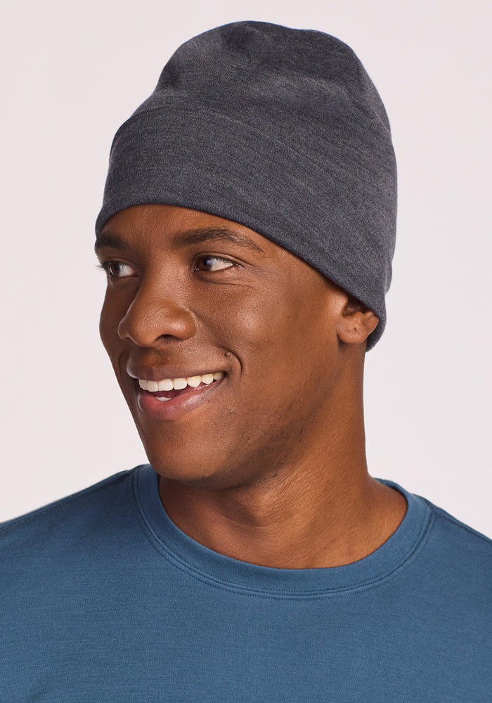 A man wearing the Woolx Journey Hat in Cinder Melange and a blue crew neck shirt smiles while looking to his left, set against a plain light background.