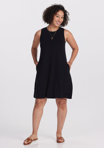 A person with curly hair smiles in a stylish Woolx Clara Dress - Black, paired with brown sandals. They have their hands in their pockets, standing against a plain white background. | Tori is 5'7", wearing a size S