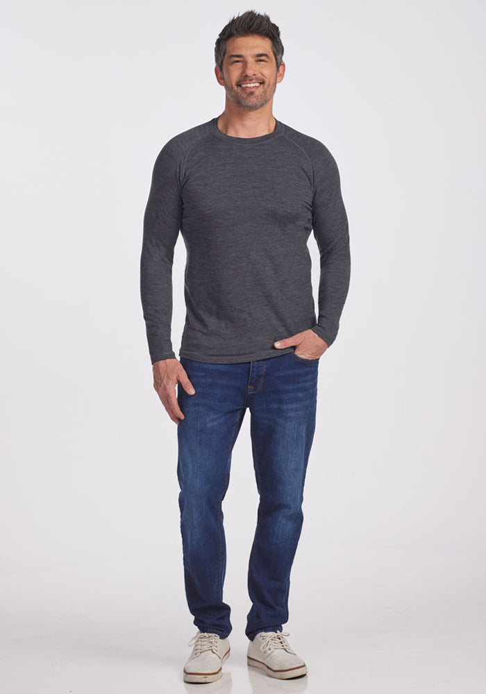 A man stands against a white background, smiling in the Woolx Essential Tee - Cinder Melange, paired with blue jeans and white sneakers. One hand in his pocket, he highlights the comfort and temperature-regulating benefits of this tee.
