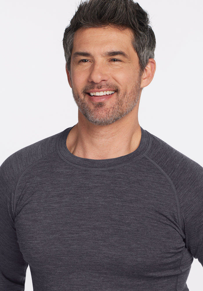 A man with short, dark hair and a trimmed beard smiles while wearing the Woolx Essential Tee in Cinder Melange, known for its temperature-regulating Merino wool. He stands against a plain white background.