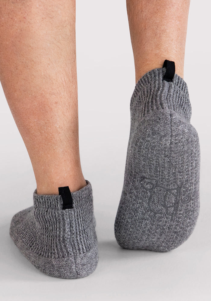 Close-up of a person's lower legs and feet wearing Woolx Mens Gripper Slippers in Gentle Grey, featuring black pull tabs and subtle sole embroidery, standing on a light-colored surface.