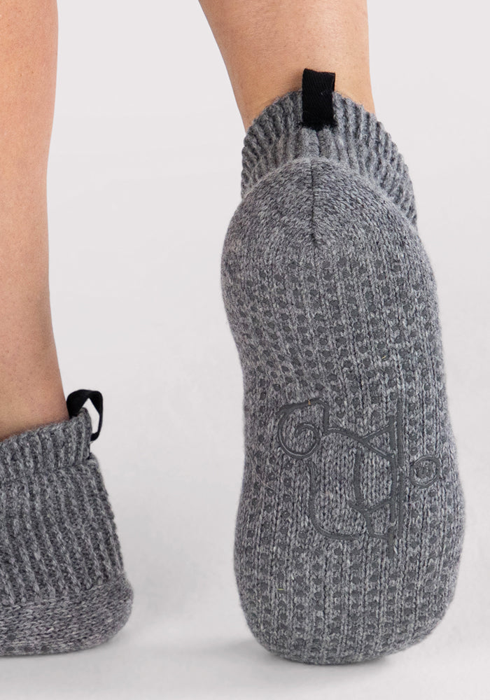 A close-up of feet wearing Woolx Womens Gripper Slippers in Gentle Grey, featuring thick ribbed knit, black pull tabs, and a subtle embroidered sole logo against a plain light background.