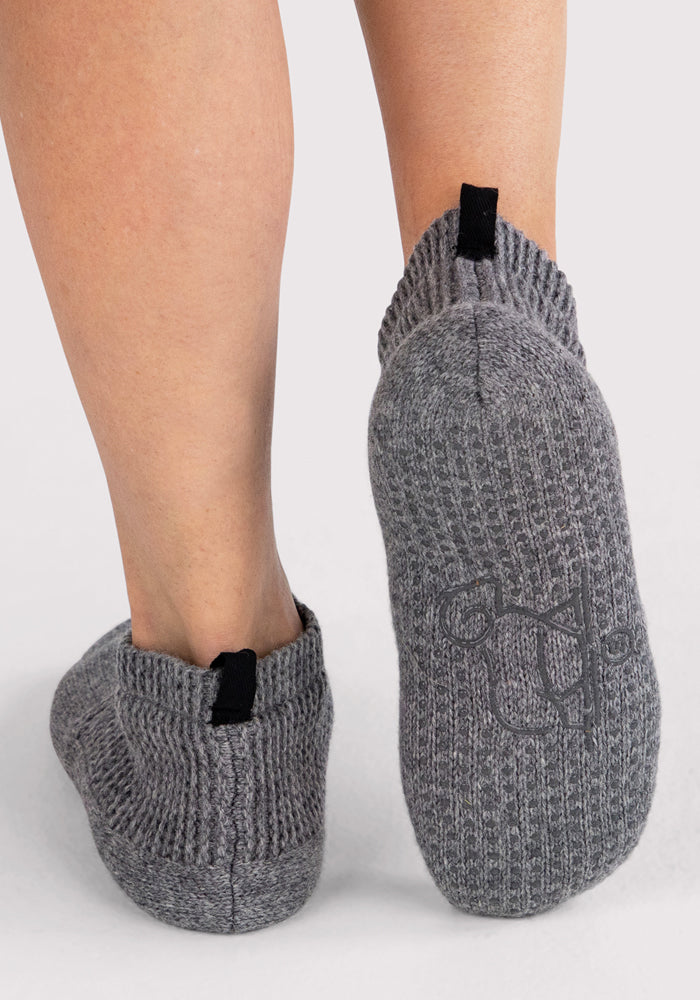 Close-up of a person wearing Woolx Womens Gripper Slippers in Gentle Grey, featuring black pull tabs and ribbed, ankle-high design. One slipper shows the decorative grip pattern on the sole. Background is plain and light-colored.
