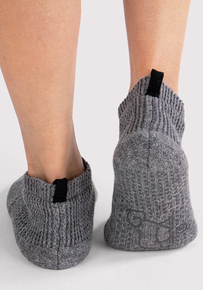 Close-up of the back of a person's lower legs wearing Woolx Womens Gripper Slippers in Gentle Grey, featuring textured fabric, black pull tabs, and non-slip grips on the soles, standing on a light neutral background.