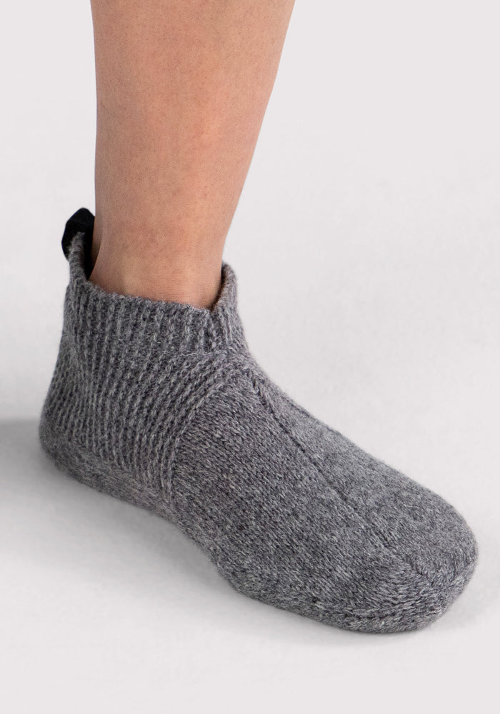 A person's lower leg and foot wear Woolx Womens Gripper Slippers in Gentle Grey, featuring a ribbed cuff and seamless design—perfect for warmth and comfort against a plain light background.