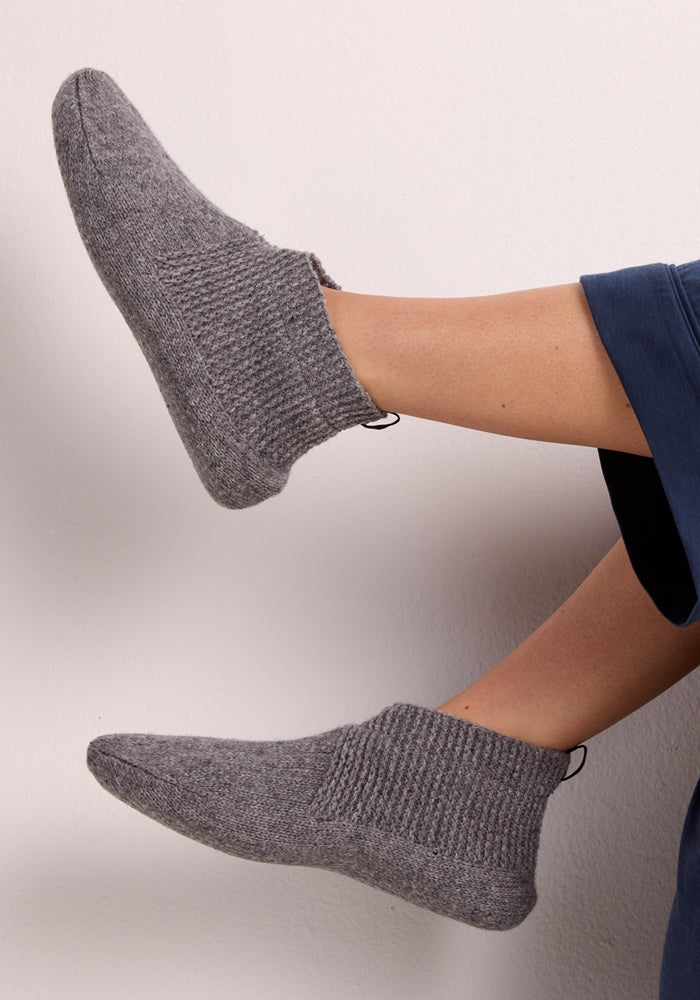 A pair of feet in Woolx Womens Gripper Slippers - Gentle Grey are crossed at the ankles against a light background, paired with dark blue pants.