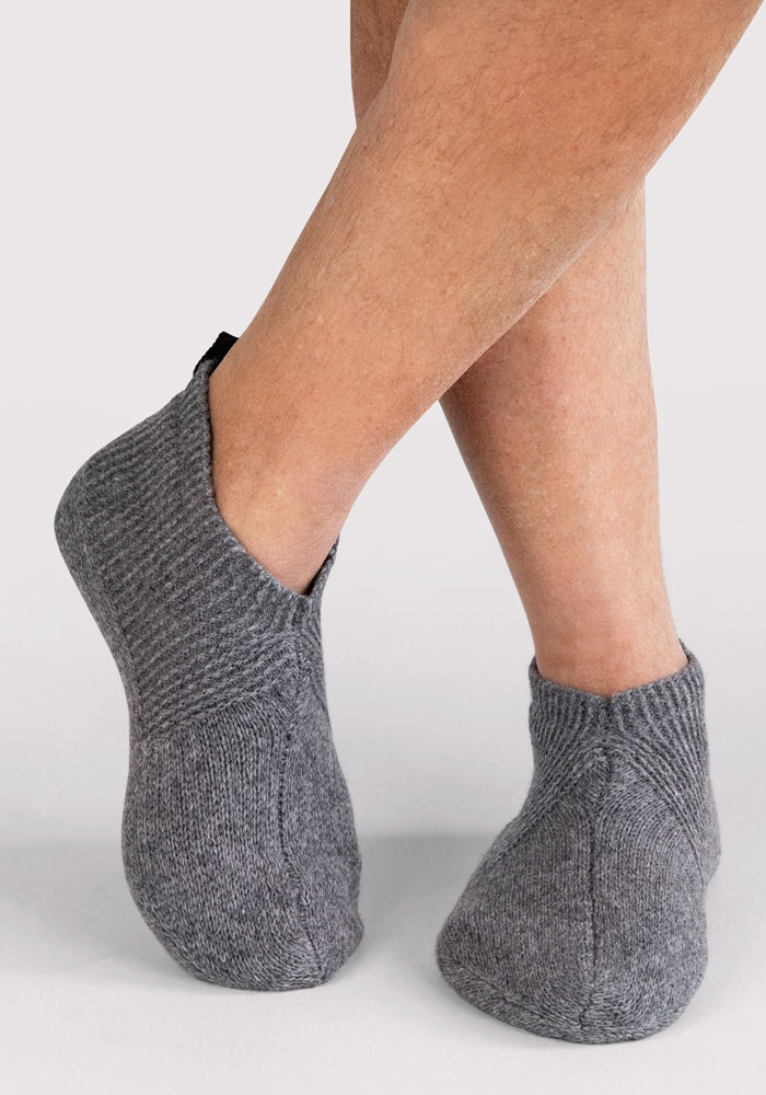 Close-up of a person's crossed legs wearing Woolx Mens Gripper Slippers in Gentle Grey, featuring thick knit fabric and gripper soles, standing on a light-colored surface against a plain background.