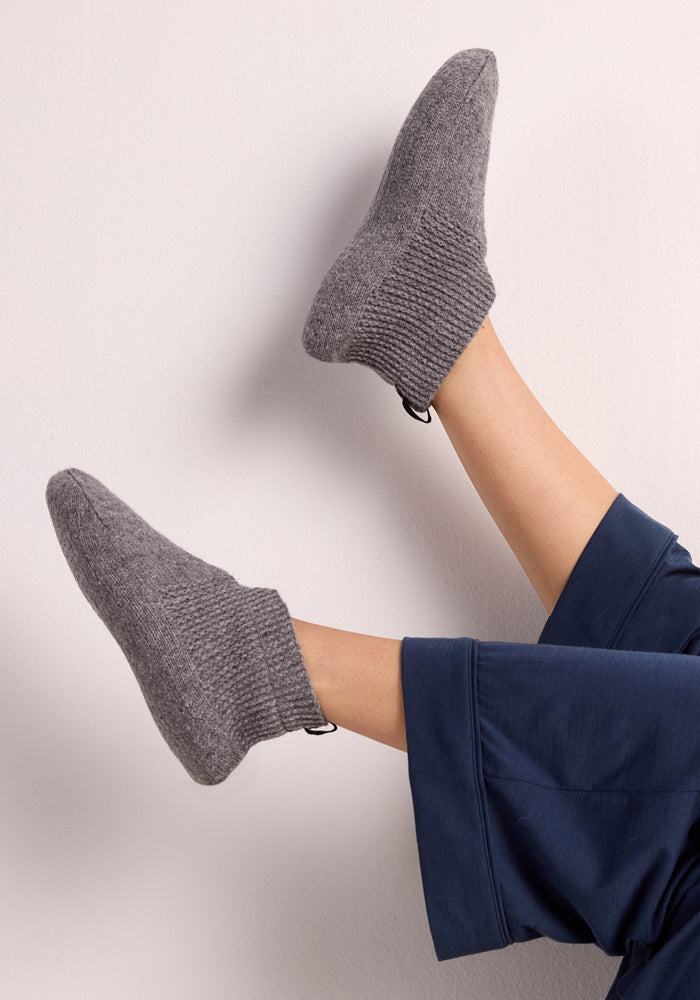 A person wearing Woolx Womens Gripper Slippers in Gentle Grey and navy blue pants has their legs raised and crossed against a neutral background.