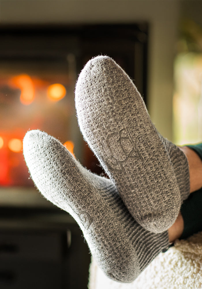 A close-up of two feet in Woolx Womens Gripper Slippers - Gentle Grey, propped by a cozy fireplace, exudes warmth and relaxation.