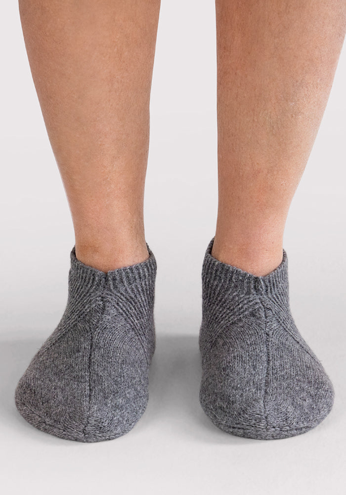 A pair of feet wears Woolx Mens Gripper Slippers in Gentle Grey, shown from the back against a plain white background.