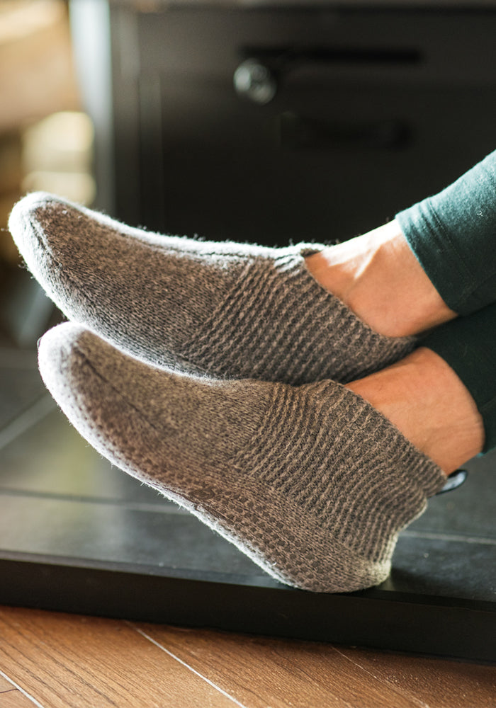 A close-up of two feet in Woolx Womens Gripper Slippers - Gentle Grey, resting on a dark surface. The wearer has on dark pants, and warm light highlights the slippers' soft, cozy texture and secure grip.